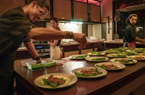 Шеф-повар посыпает гарниром тарелку с едой, а пара других поваров наблюдает за этим. Стол заполнен блюдами, наполненными едой на кухне; Устойчивое Пуэрто-Рико