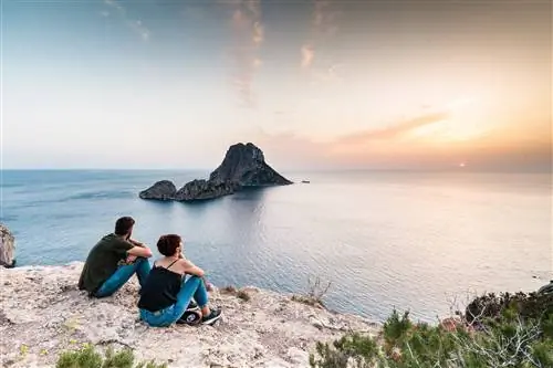 Два человека сидят на скалистом выступе и смотрят в море на островок, за которым садится солнце.