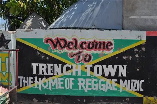 Бетонная стена со словами Trench Town The Home of Reggae Muzic нарисована поверх картины с изображением зелено-черно-золотого флага Ямайки.