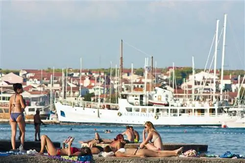 Люди в купальниках загорают на пирсе на Адриатическом побережье с парусниками, кораблями и домами на расстоянии позади них