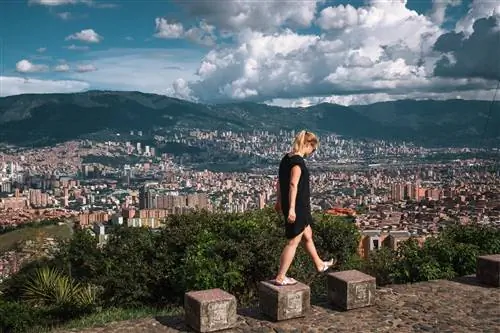 Вид сбоку женщины, идущей по каменным сиденьям со зданиями на заднем плане против облачного неба