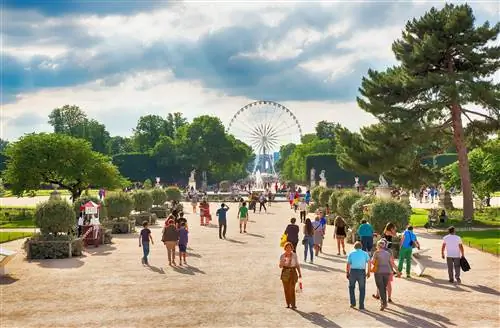 Отдельные группы людей идут по широкой тропинке через сад к большому колесу обозрения вдалеке.