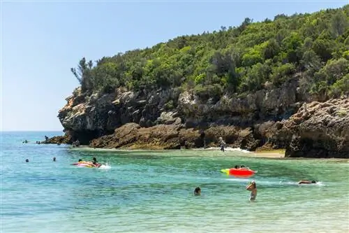 Зеленый склон холма над скалами, которые нависают над пловцами в воде в Прайя-дос-Галапиньюс, Португалия, Европа