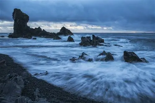 Пляж с черным песком и шумным прибоем. В море возвышаются несколько огромных скалистых утесов.