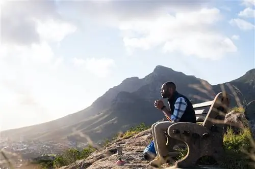 Вид сбоку на туриста-мужчину, сидящего на скамейке, выпивающего и смотрящего на Столовую гору.