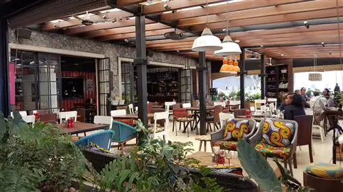 Интерьер ресторана Nyama Mama в Уэстлендсе. Он залит солнцем, с плетеными стульями разных цветов. Стальные балки поддерживают деревянные эстакады