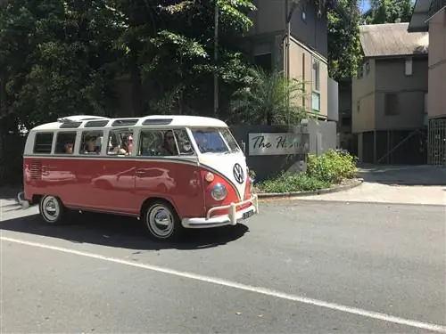Ретро-фургон VW Kombi с ярко-красным днищем и белым верхом (которые сходятся в точке на переднем бампере) едет по обсаженной деревьями улице в Нусе; вдоль борта фургона и по краю крыши есть множество отдельных окон, а середина крыши убирается, а ткань складывается ближе к задней части.