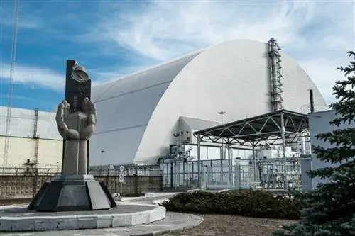На переднем плане бетонный памятник Чернобыльской катастрофе, изображающий две руки, сложенные вместе вокруг миниатюры реактора № 4; за мемориалом находится огромная серая арка конструкции Нового безопасного конфайнмента, которая окружает остатки реактора 4.