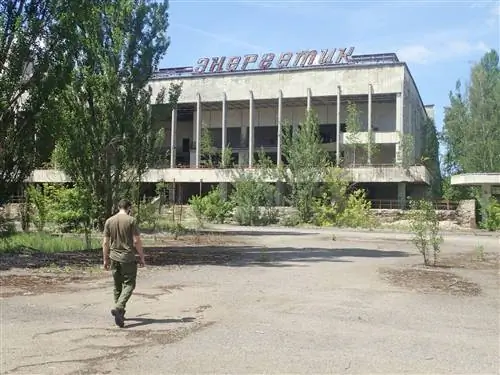 Гид идет по асфальту к заброшенному Дворцу культуры в Припяти; Деревья растут перед бетонной конструкцией с колоннами и внутри нее, на крыше которой есть большая вывеска украинской вязью.