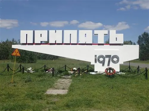 Бетонный знак украинским шрифтом, который переводится как Припять 1970 года, когда город был основан; под ним расставлены венки и цветы, а рядом стоит табличка с предупреждением о радиации.
