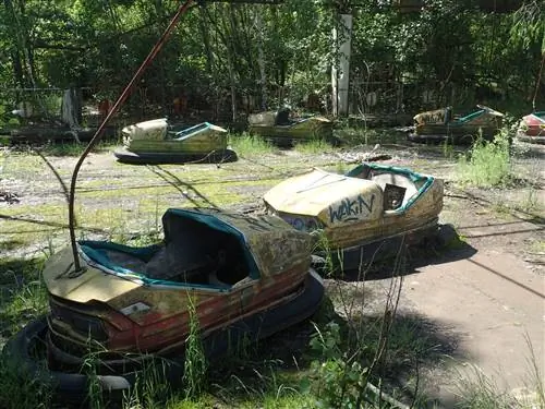 Ветхие желтые и синие автомобили Dodgem в парке развлечений Припять, между ними и деревьями на заднем плане растут сорняки.