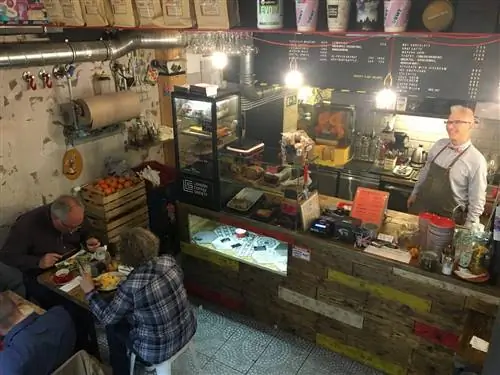 Глядя вниз на переднюю стойку London Coffee Society из мезонина, мужчина за стойкой широко улыбается, пока клиенты обедают на своем будапештском позднем завтраке.