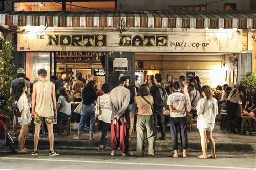 Большая толпа людей собирается в баре North Jazz Co-Op, чтобы послушать живую музыку. Толпа выплескивается из бара на тротуар снаружи.