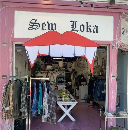 Внешний вид магазина Sew Loka. С витрины свисает вывеска в виде пары губ с клыками. Сбоку видна вешалка с одеждой и можно заглянуть внутрь магазина.