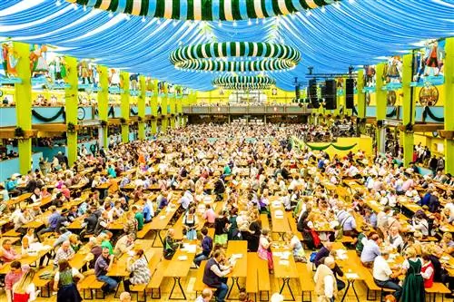 Люди в пивной палатке Spaten на Октоберфесте в Мюнхене. Огромный шатер, вмещающий сотни столов, с ярким голубым потолком и полосатым декором.