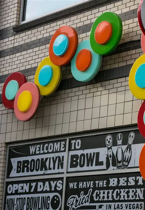 Вывески на внешней стороне Brooklyn Bowl в Лас-Вегасе имеют белый текст на черном фоне и случайный набор цветных дисков, которые напоминают пластинки над головой.