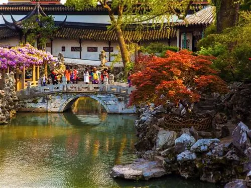 Пруд с небольшим каменным мостиком и крытой дорожкой на заднем плане в обрамлении деревьев с красной листвой. Классическая китайская ландшафтная архитектура, отмеченная павильонами, крытыми дорожками, скальными элементами и прудами, была разработана в Сучжоу.