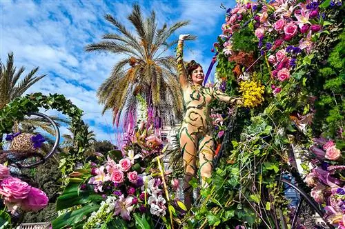 Женщина в обнаженном костюме машет толпе с парадной платформы, усыпанной цветами.