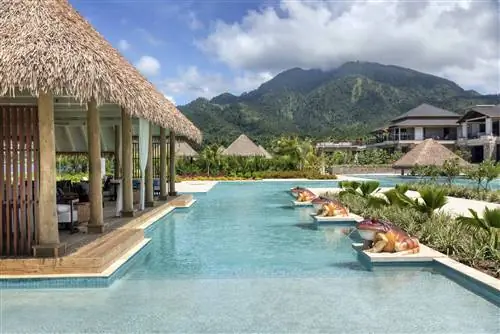 Внешний вид бассейна курорта Cabrits Resort с тремя большими статуями лягушек, плюющихся водой в большой бассейн. Есть большая открытая общая зона с соломенной крышей, заполненная стульями и столами. Прядь облаков покрывает вершину большой зеленой горы вдалеке.