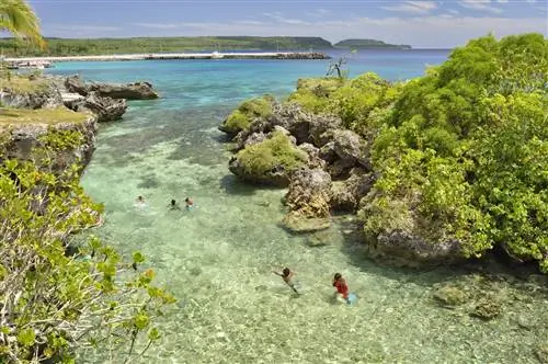 Лагуна на острове Маре, Новая Каледония. В чистой воде плещутся несколько человек. По обеим сторонам лагуны расположены громадные серые скалы, увенчанные яркими зелеными растениями и мхом.