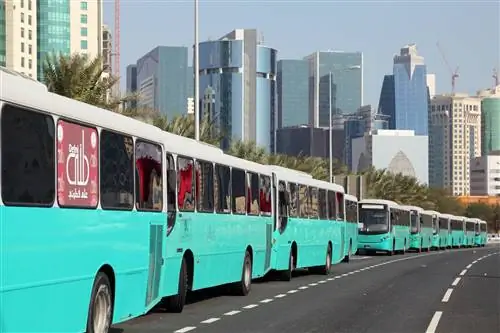 Несколько бирюзово-синих автобусов выстроились в линию на дороге с башнями в Дохе, Катар.