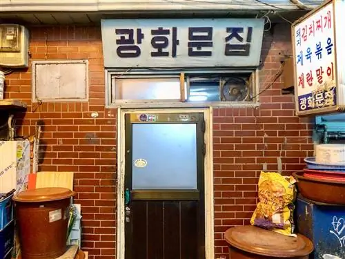 Дверь Gwanghwamun Jip в Сеуле с табличкой на корейском языке над ней и несколькими мусорными баками по бокам вдоль кирпичной стены.
