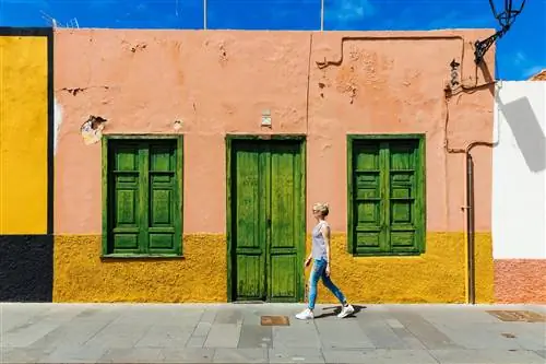Белая женщина проходит мимо одноэтажного розово-желтого дома с зелеными ставнями в Пуэрто-де-ла-Крус, Тенерифе.