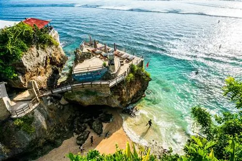 Серфер выходит к океану на пляже Сулубан, рядом с отвесной скалой с гостиной на вершине.