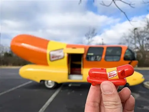 Рука держит игрушечный свисток Weenie Whistle на фоне Винермобиля Оскара Майера.