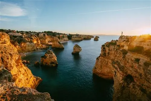 Скалистая береговая линия Португалии в Прайя-да-Маринья залита теплым солнечным светом; груды скал поднимаются из спокойного моря, и видна фигура, стоящая на вершине утеса.