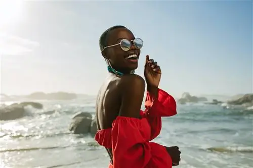 Черная женщина в красном платье и солнцезащитных очках оглядывается через плечо и смеется на пляже.