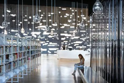 Женщина сидит в библиотеке Espacio De Las Artes на Тенерифе. Стеклянные скульптуры, похожие на капли дождя, свисают с потолка, а на заднем плане виден современный белый письменный стол.