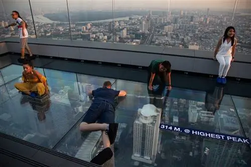 Посетители сидят и лежат на стеклянном полу на крыше здания King Power Mahanakhon в Бангкоке, в то время как огни городского горизонта сверкают где-то внизу.