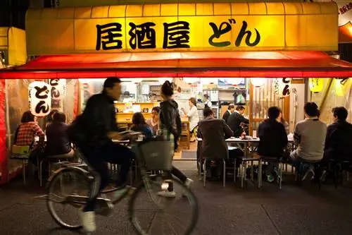 Мужчина проезжает на велосипеде мимо кафе в Токио, которое открыто для улицы, и посетители и пьющие видны под его навесом и висящими фонарями.