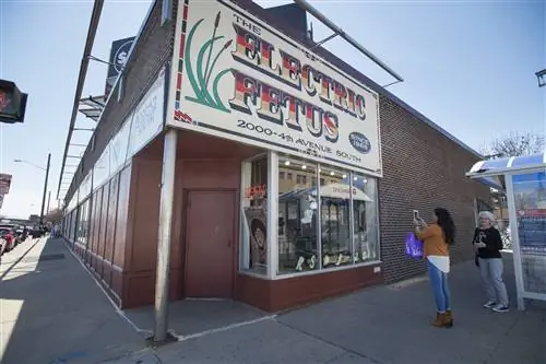 Женщина в синих узких джинсах, коричневых ботинках, белом топе и свитере верблюжьего цвета фотографирует на свой телефон здание Electric Fetus, кирпичную витрину с большим лиловым, оранжевым и коричневым шрифтом в стиле 70-х. и иллюстрация кошачьих хвостов с развевающимися листьями.