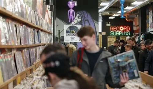 Покупатели разбирают корзины с альбомами на Shuga Records. В центре изображения молодой человек с рыжими волосами в сером худи с красным рюкзаком, держащий в руках синий альбом. В центре внимания позади него фиолетовая фреска, фиолетовая надувная игрушка-инопланетянин и неоновая вывеска красного и зеленого цветов с надписью Record Surplus, где O в Record выглядит как альбом