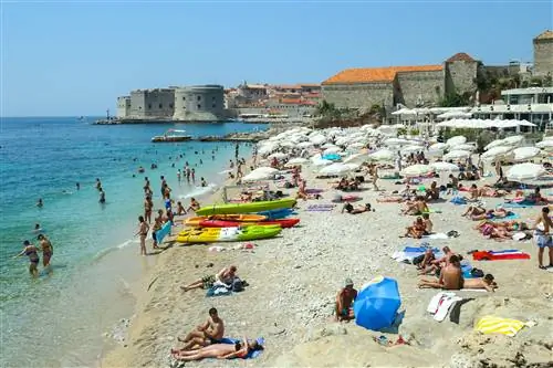Люди купаются и загорают на морском пляже Банье на фоне старого города Дубровника.