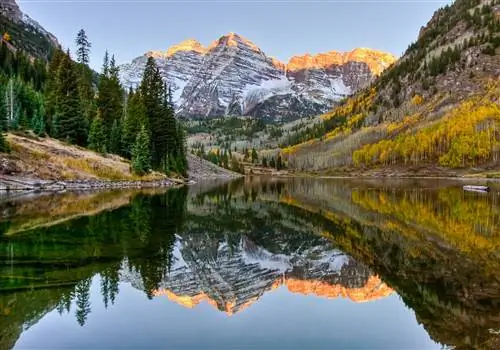 Восход солнца падает на заснеженные пики Maroon Bells, отражаясь в озере внизу, вместе с деревьями осины в их осенней листве, отображающей золотые цвета.