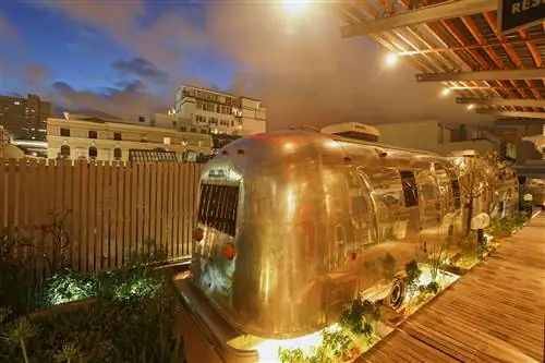 Блестящие серебристые трейлеры Airstream сияют золотым светом благодаря теплому деревянному забору, настилу и потолку над тщательно ухоженными площадками, где они припаркованы в отеле Grand Daddy в Кейптауне. На два трейлера, припаркованных в ряд, светят огни, падающие на них с земли и падающие с нависающей крыши. За ними и забором белые многоэтажки намекают на окружающий район.