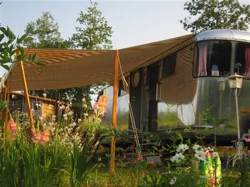 Серебристый кемпер Airstream с тентом в бело-оранжевую полоску, выдвинутым с левой стороны, стоит в саду с полевыми цветами и коралловыми гладиолусами. На заднем плане деревянная баня добавляет деревенского очарования.