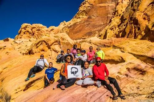 Группа чернокожих туристов позирует с небольшим баннером Outdoor Afro на скалах песочного цвета.