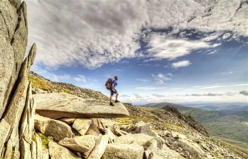 Мужчина в туристическом снаряжении с рюкзаком стоит на скалистом выступе с холмистой местностью вдалеке.
