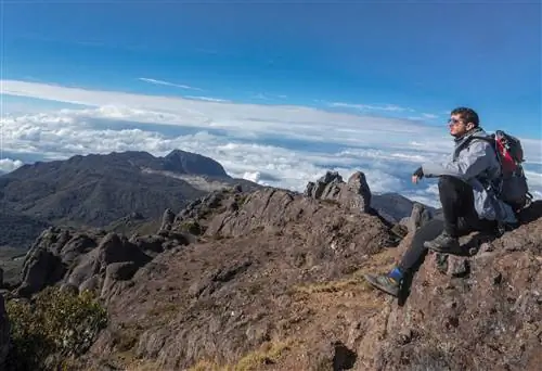 Солнечным утром в Национальном парке Чиррипо, Коста-Рика, Центральная Америка, мужчина сидит на скале и смотрит на вершины и облака.