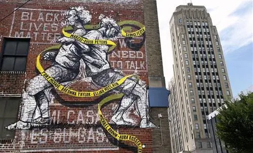 Фреска на здании с кирпичными стенами, изображающая двух человек спиной к спине, стоящих со сцепленными руками. Вокруг них вьется желтая лента с именами чернокожих жертв полицейских расстрелов.