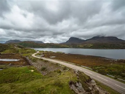 Узкая дорога вьется вдоль водоема в Шотландском нагорье; впереди на невысоких холмах висят серые тучи.