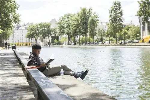 Мужчина сидит на берегу канала Сен-Мартен в Париже и читает книгу в солнечный день.