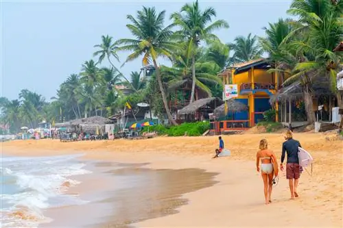 Женщина и мужчина с досками для серфинга идут по пляжу с барами, пляжными лачугами и пальмами.