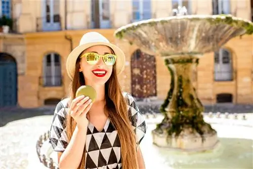 Женщина в очках стоит рядом с фонтаном в городе Экс-ан-Прованс, улыбается и держит круглый фисташковый макарон.