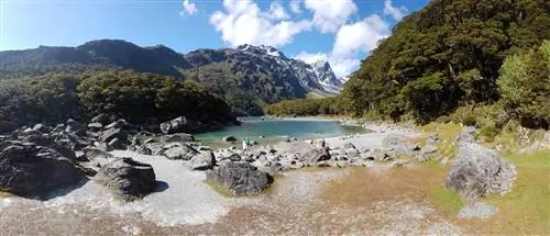панорамный снимок озера Маккензи на Южном острове Новой Зеландии в солнечный день. У самой кромки воды стоят и лежат на небольшом каменистом пляже несколько человек. Голубая вода окружена скалистыми, заснеженными горами и пышной зеленью.