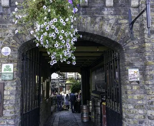 Кадр через арочный каменный проем во двор паба, где собираются люди.